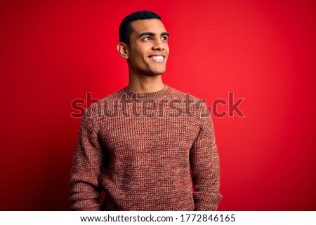 Young handsome african american man wearing casual sweater standing over red background looking away to side with smile on face, natural expression. Laughing confident.