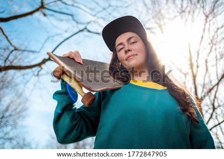 A young caucasian hipster smiling teen poses with a skateboard over her shoulder. In the background, blue sky, trees and sun. Bottom view. Close up. Concept of sports lifestyle and street culture