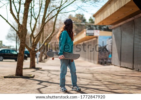 A young Caucasian hipster woman walks down the street with a skateboard in her hands and turns back. In the background, an alley. Concept of sports lifestyle and street culture
