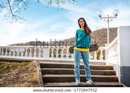 A young Caucasian woman holding a skateboard behind her back is standing on the steps. In the background, blue sky and trees. Bottom view. Concept of sports lifestyle and street culture