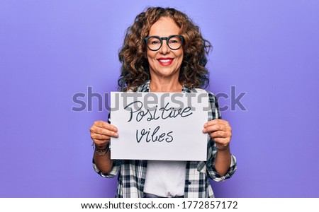 Middle age woman asking for optimistic attitude holding paper with positive vibes message looking positive and happy standing and smiling with a confident smile showing teeth