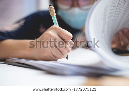 An asian lady wearing a surgical mask signs some documents with a ballpen. New normal concept.