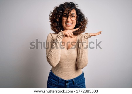 Young beautiful curly arab woman wearing casual t-shirt and glasses over white background amazed and smiling to the camera while presenting with hand and pointing with finger.