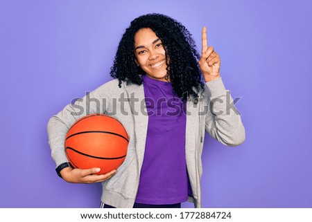 African american curly sportswoman doing sport holding basketball ball over purple background surprised with an idea or question pointing finger with happy face, number one