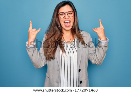 Young hispanic business woman wearing glasses standing over blue isolated background shouting with crazy expression doing rock symbol with hands up. Music star. Heavy music concept.