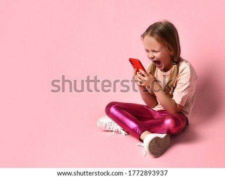 Blonde child in t-shirt, leggings and sneakers. She is screaming while looking at screen of her red smartphone while sitting on floor with crossed legs against pink background. Close up, copy space