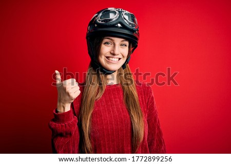 Young beautiful redhead motocyclist woman wearing moto helmet over red background doing happy thumbs up gesture with hand. Approving expression looking at the camera showing success.