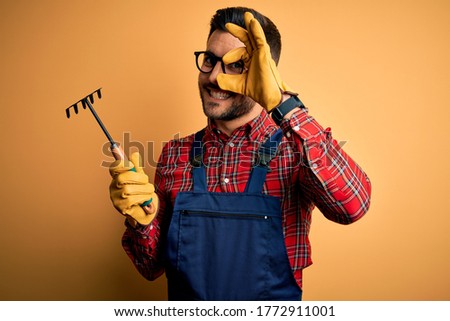 Young gardener man wearing working apron using gloves and tool over yellow background with happy face smiling doing ok sign with hand on eye looking through fingers