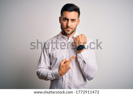 Young handsome man wearing elegant shirt standing over isolated white background In hurry pointing to watch time, impatience, looking at the camera with relaxed expression