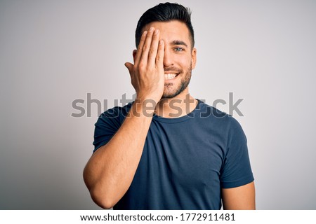 Young handsome man wearing casual t-shirt standing over isolated white background covering one eye with hand, confident smile on face and surprise emotion. Royalty-Free Stock Photo #1772911481