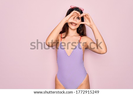 Young beautiful fashion girl wearing swimwear swimsuit and sunglasses over pink background Doing heart shape with hand and fingers smiling looking through sign