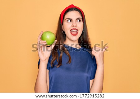 Young beautiful girl eating fresh organic healthy green apple over yellow background pointing and showing with thumb up to the side with happy face smiling