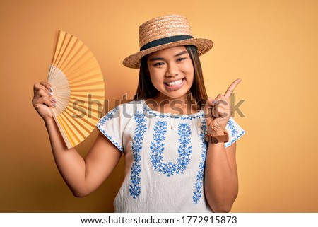 Young asian tourist girl on vacation wearing summer hat using hand fan over yellow background very happy pointing with hand and finger to the side