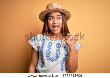 Young beautiful asian girl wearing casual t-shirt and hat standing over yellow background smiling with happy face looking and pointing to the side with thumb up.