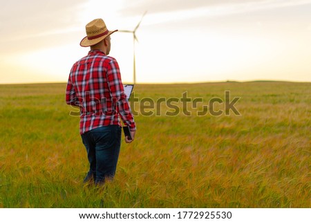 Young farmer standing in filed of wheat with tablet in his hands. Looking in wind mills