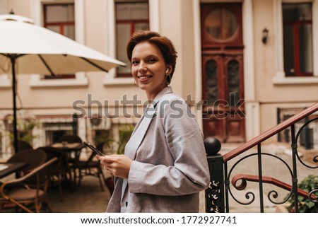 Cool woman looks into camera and holds tablet. Cheerful dark-haired lady in grey stylish jacket widely smiles and poses in house yard