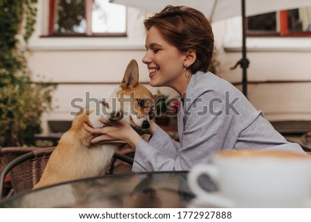 Joyful woman in grey outfit laughing and playing with corgi in street cafe. Short-haired lady in jacket smiles and poses with dog