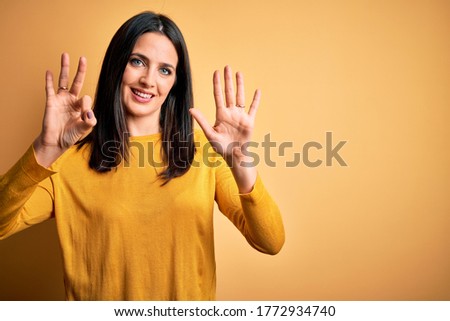 Young brunette woman with blue eyes wearing casual sweater over yellow background showing and pointing up with fingers number eight while smiling confident and happy.