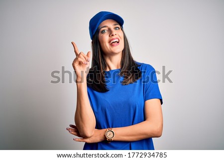 Young delivery woman with blue eyes wearing cap standing over blue background with a big smile on face, pointing with hand and finger to the side looking at the camera.