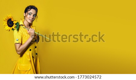The girl in a yellow dress holds a large sunflower and smiles playfully.