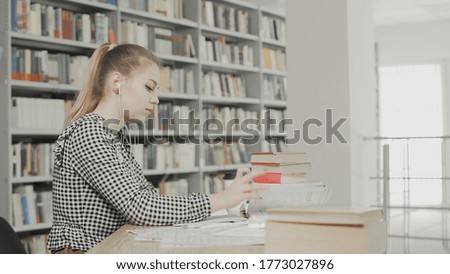 Concentrated girl student with headphones preparing for examination and writing notes while sitting at table at university library