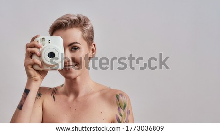Portrait of a beautiful tattooed woman with pierced nose and short hair using instant photo camera, standing isolated over grey background. People and tehnology concept. Web Banner