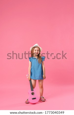 cheerful little Caucasian girl of 4 years old in a short denim dress and a straw hat poses in the Studio with a pink ukulele guitar. A child plays a musical instrument and sings on a pink background.