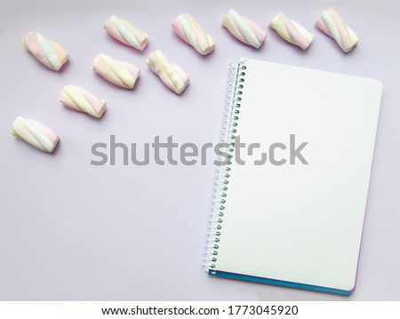 Close up picture of notebook on right side and marshmallow on the top laying on light lavender background. Copy space concept with empty space for text in pastel tones. Flat lay composition.