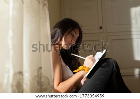 Young hispanic woman writing in a black notebook, thoughtful girl writing in her journal in her room at sunset. young man near the window concentrated studying at home. Royalty-Free Stock Photo #1773054959