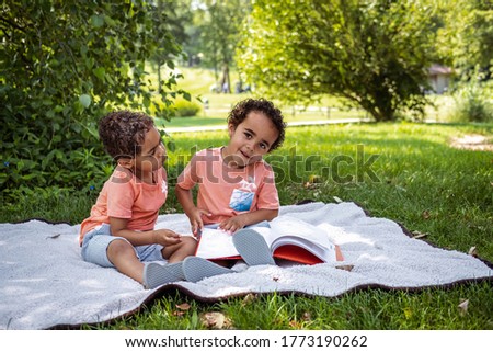 African-American three year old twin boys sit on blanket in park with book in front of them; one boy is looking into camera