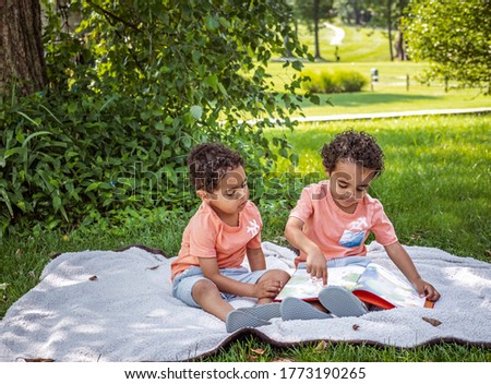 Three year old African-American twin boys  looking sitting on blanket in park looking at book in front of them