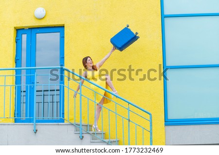 A happy red-haired woman in a yellow dress is walking down the stairs and joyfully swing a blue suitcase. The girl is preparing for the journey. Summer vacation concept.