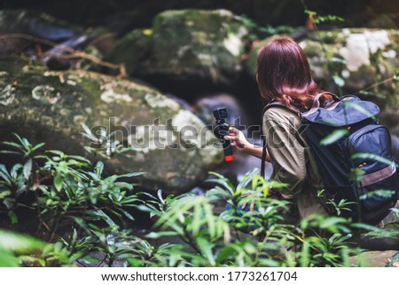 A female tourist with backpack shooting video of the waterfall in the forest by an action camera