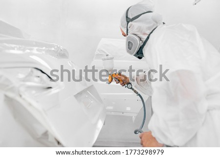 Worker in protective uniform spraying car body part in white paint. Royalty-Free Stock Photo #1773298979