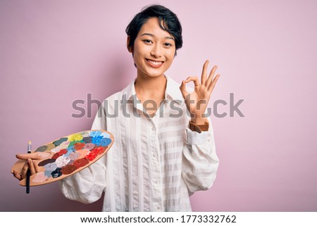 Young beautiful chinese artist woman painting using paintbrush and palette with colors doing ok sign with fingers, excellent symbol