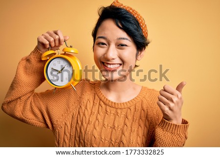 Young beautiful chinese woman holding vintage alarm clock over isolated yellow background happy with big smile doing ok sign, thumb up with fingers, excellent sign