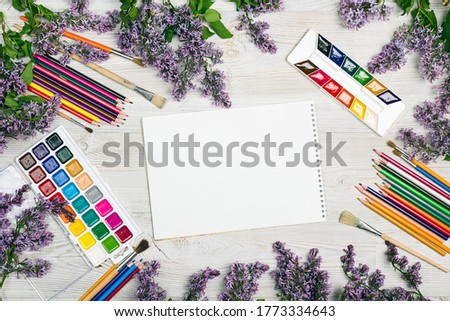 Workspace with empty sketchbook, watercolor, brushes, pencils and lilac flowers on wooden background. Flat lay, top view.