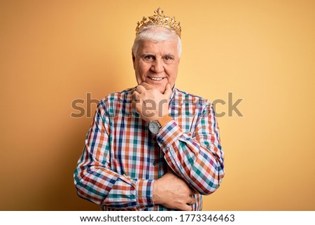 Senior handsome hoary man wearing golden crown of king over isolated yellow background looking confident at the camera smiling with crossed arms and hand raised on chin. Thinking positive.