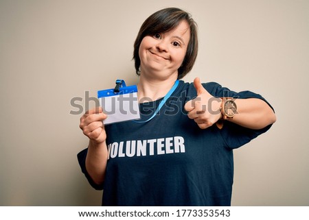 Young down syndrome volunteer woman wearing social care charity t-shirt showing identification happy with big smile doing ok sign, thumb up with fingers, excellent sign