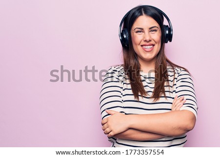 Young beautiful brunette woman listening to music using headphones over pink background happy face smiling with crossed arms looking at the camera. Positive person.