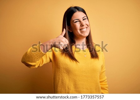 Young beautiful woman wearing casual sweater over yellow isolated background doing happy thumbs up gesture with hand. Approving expression looking at the camera showing success.