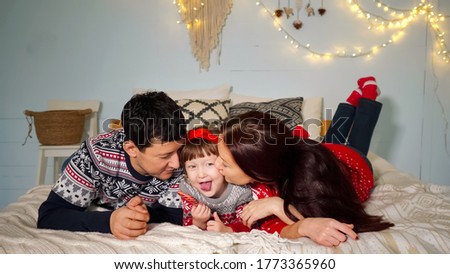 positive parents lie on cozy bed and kiss cute daughter with candy posing at family Christmas closeup