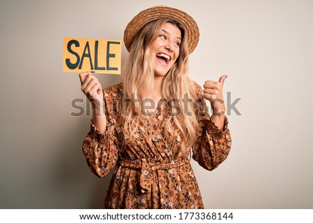 Young beautiful blonde woman holding sale banner over isolated white background pointing and showing with thumb up to the side with happy face smiling