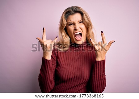 Young beautiful blonde woman wearing casual sweater over isolated pink background shouting with crazy expression doing rock symbol with hands up. Music star. Heavy concept.