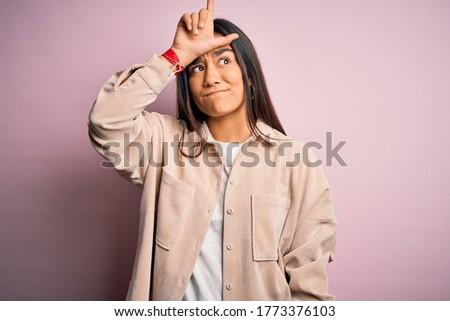 Young beautiful asian woman wearing casual shirt standing over pink background making fun of people with fingers on forehead doing loser gesture mocking and insulting.