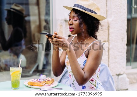 African american beautiful woman in straw hat, summer dress, taking photo on her waffles in the coffee shop, outdoor and prepared to upload to a social app.