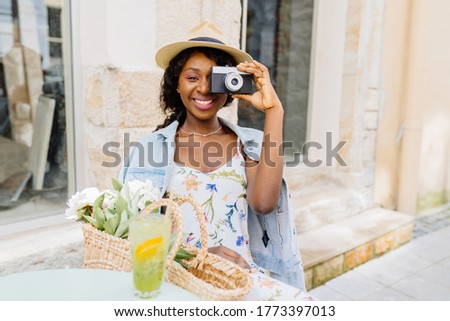 Outdoor summer lifestyle portrait of pretty excited african woman having fun in the city. Female photographer in straw hat, summer dress and jean jacket making pictures with old retro camera.