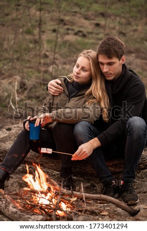 Beautiful guy and girl relax in nature, sit by the fire and roast marshmallows, active rest, love, romance, married couple