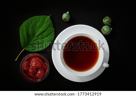 White cup with tea, strawberry jam, poppy boxes and hydrangea leaf on dark background, top view, still life