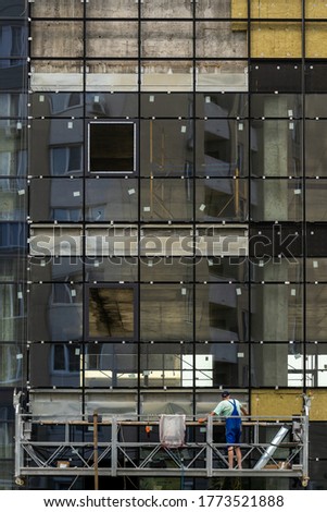 Concept of unfinished skyscraper. Vertical photo of building under construction with large glass windows and professional worker man in special scaffolding equipment